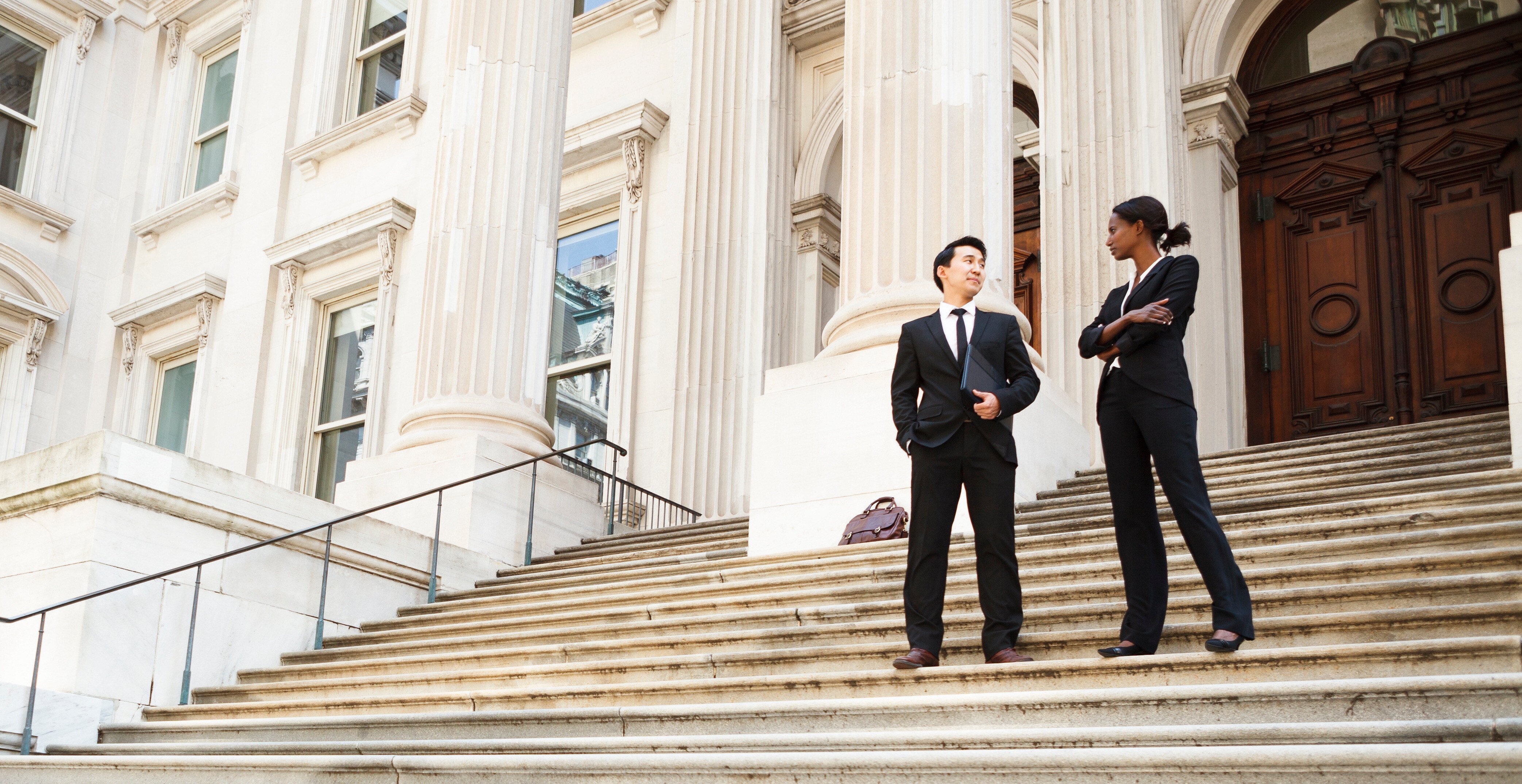 Two professionals in suits stand on the stairs of a large, official-looking building with large columns.