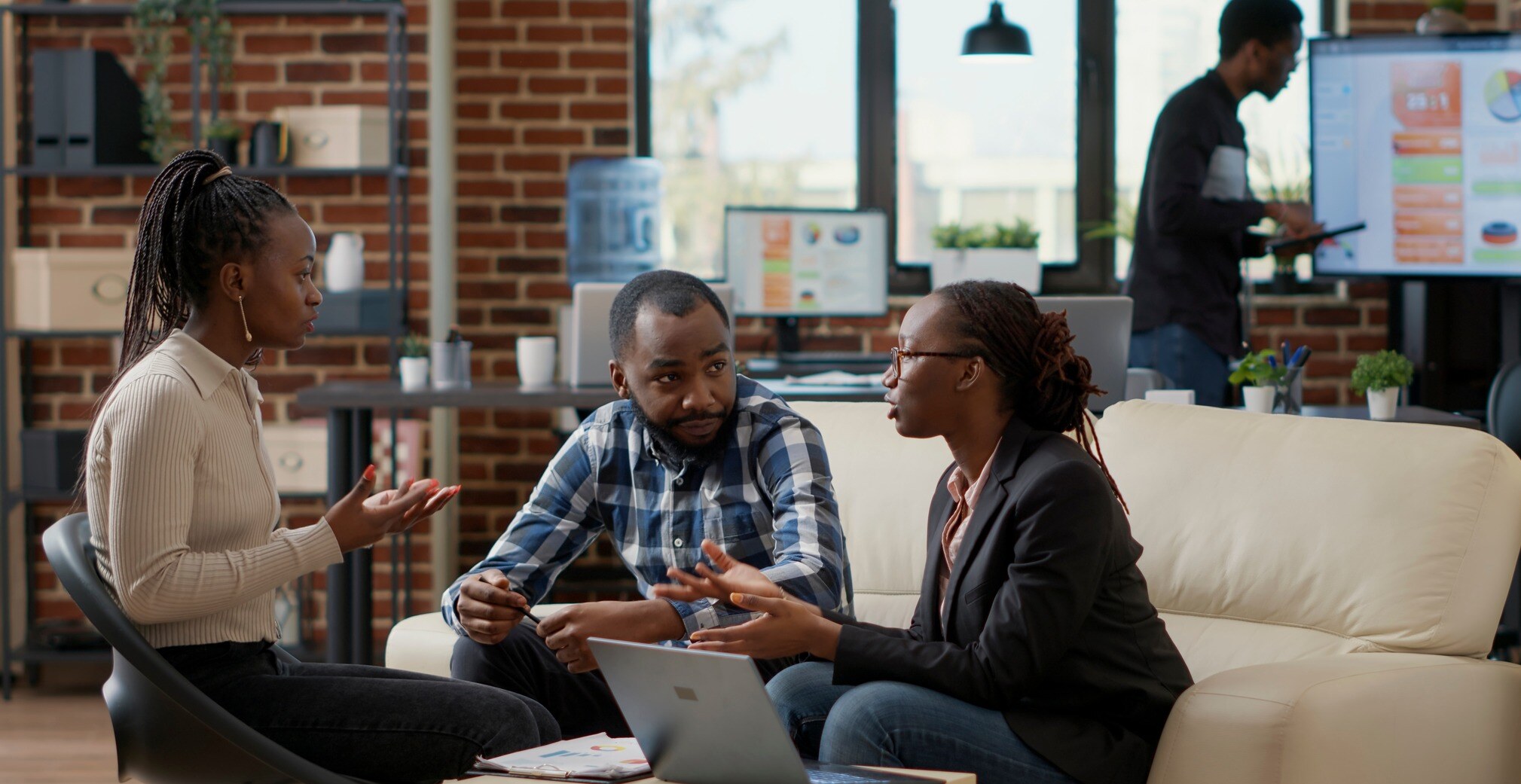 Three colleagues discussing in a modern office with laptops, while another person works in the background.