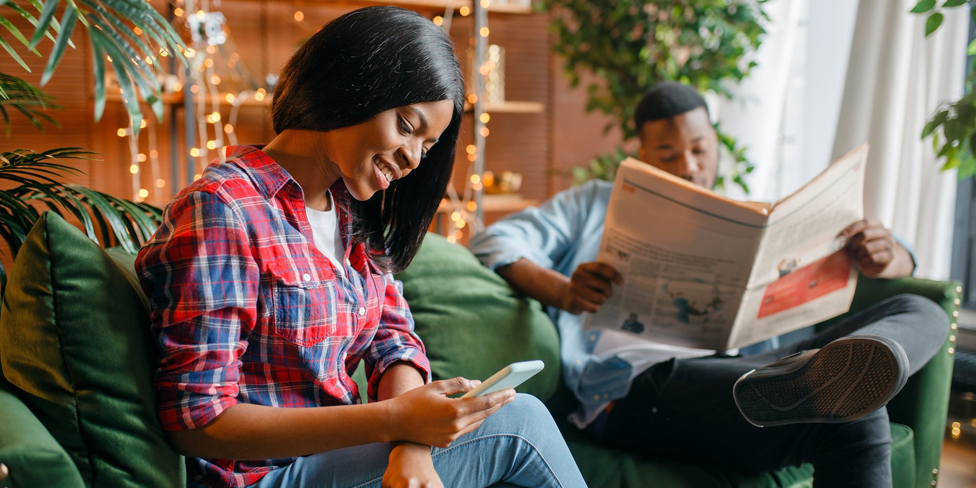 A man and a woman sit in a living room. She's on her phone, he's reading a newspaper.