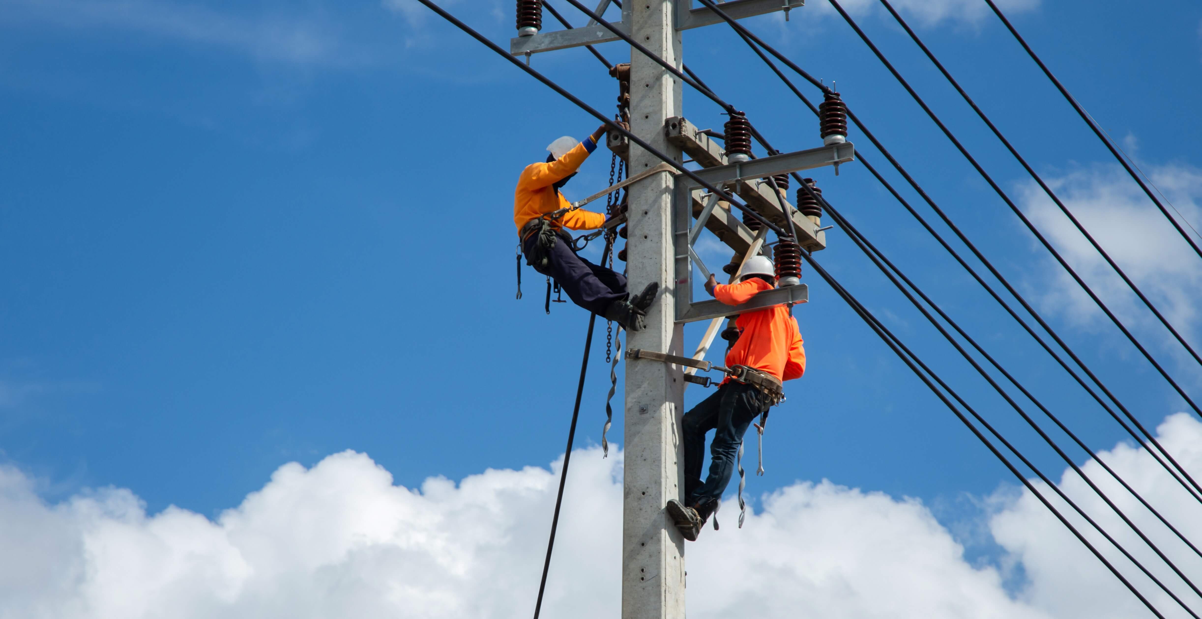 Two electricians work on a power line while tethered to a tall utility pole.