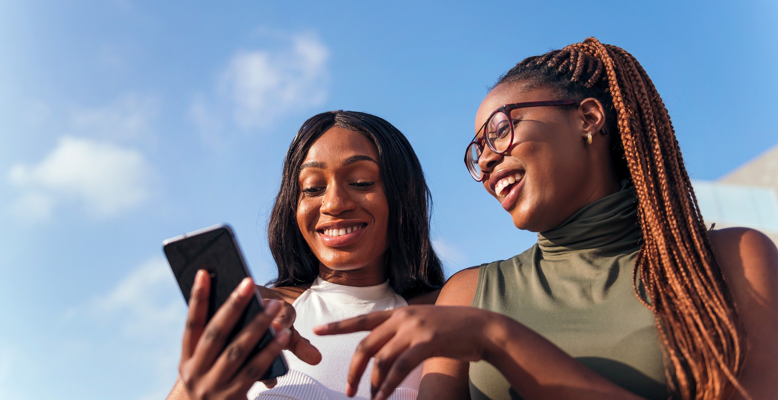 Two young adult women look at a smartphone screen while smiling.