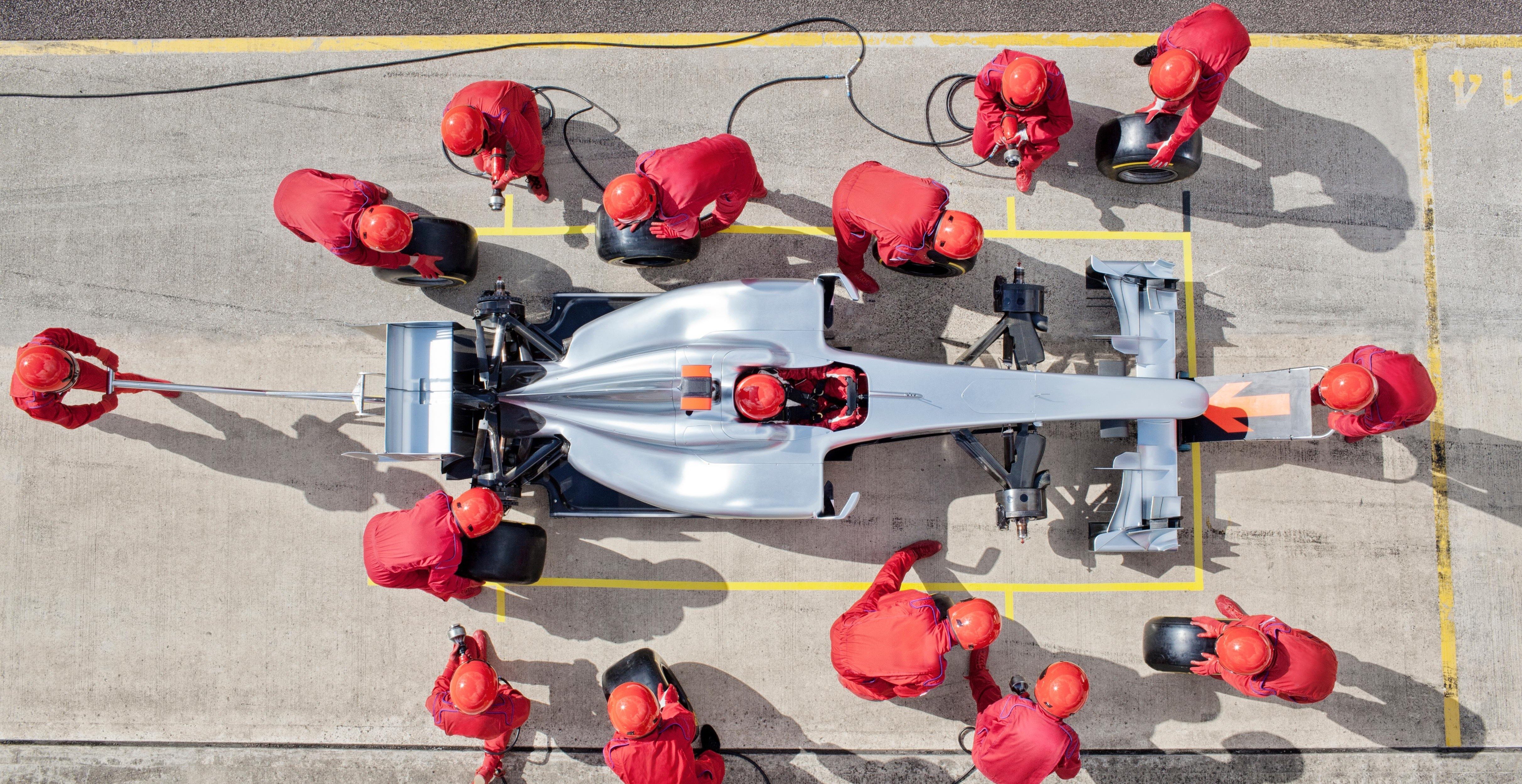 A pit crew wearing all red work together to change the tires on a silver racing car. 