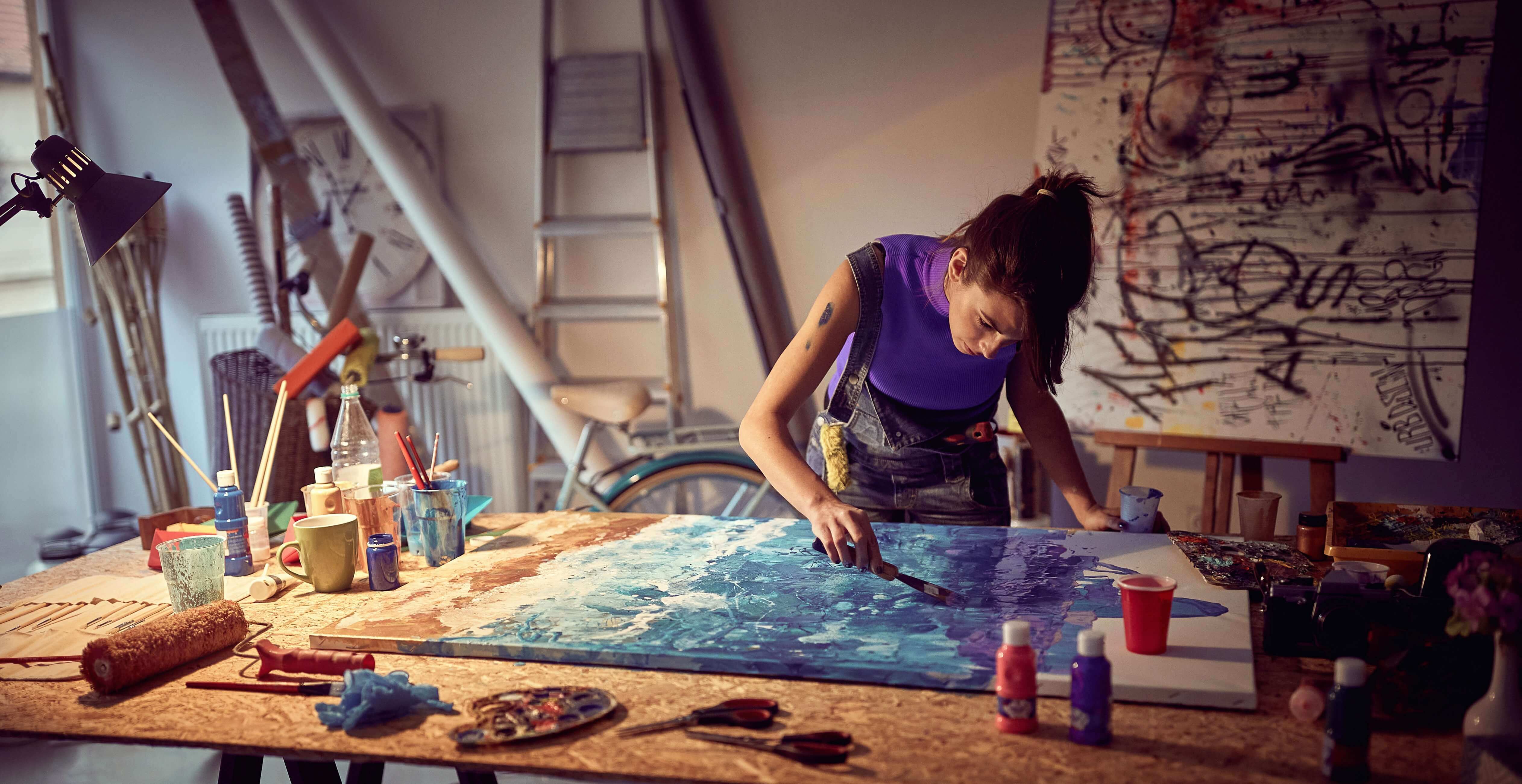 A woman wearing a paint-stained apron bends over a table to paint a large blue canvas in her artist studio.