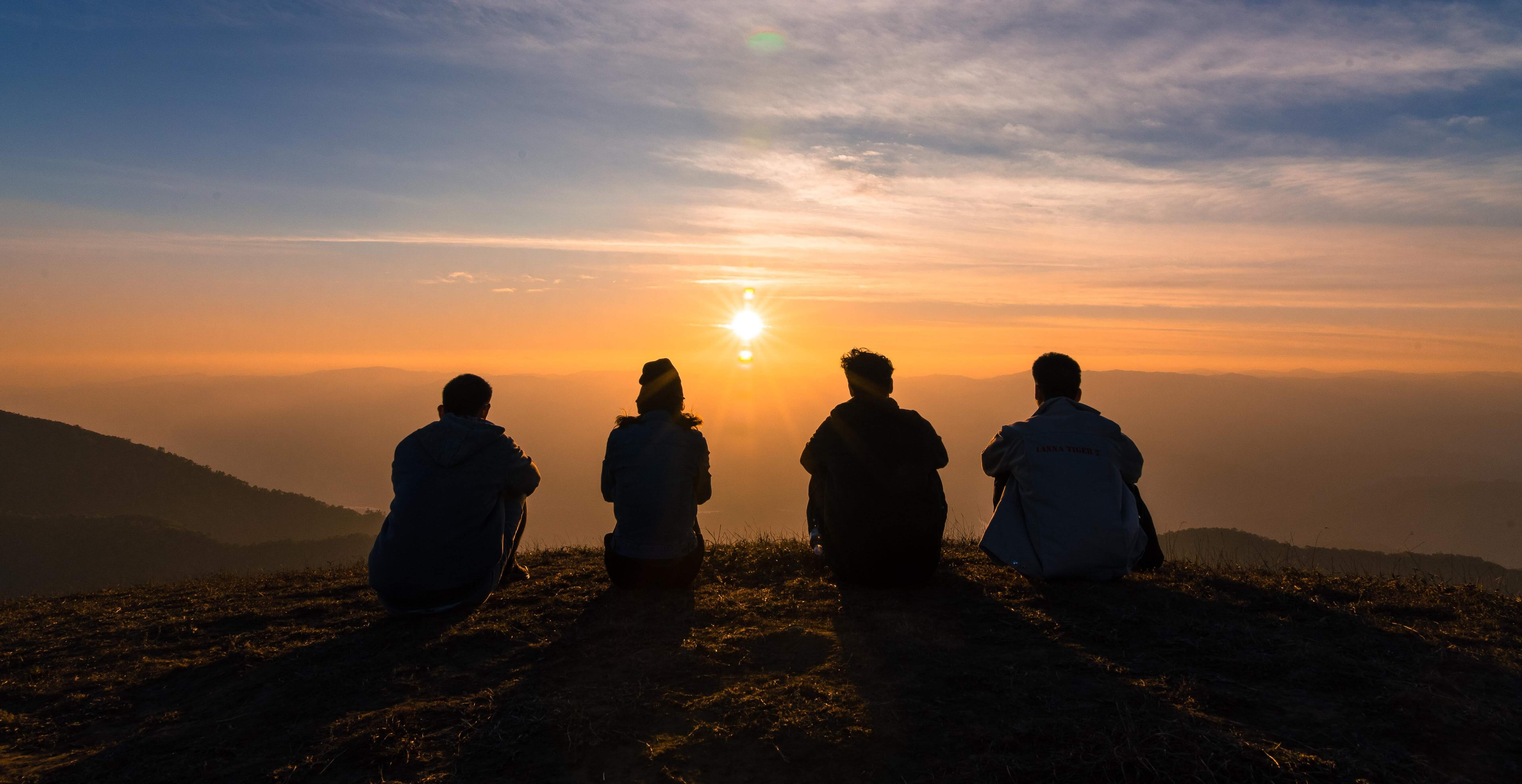 Four silhouetted people sit on a mountaintop while looking at a sunset.