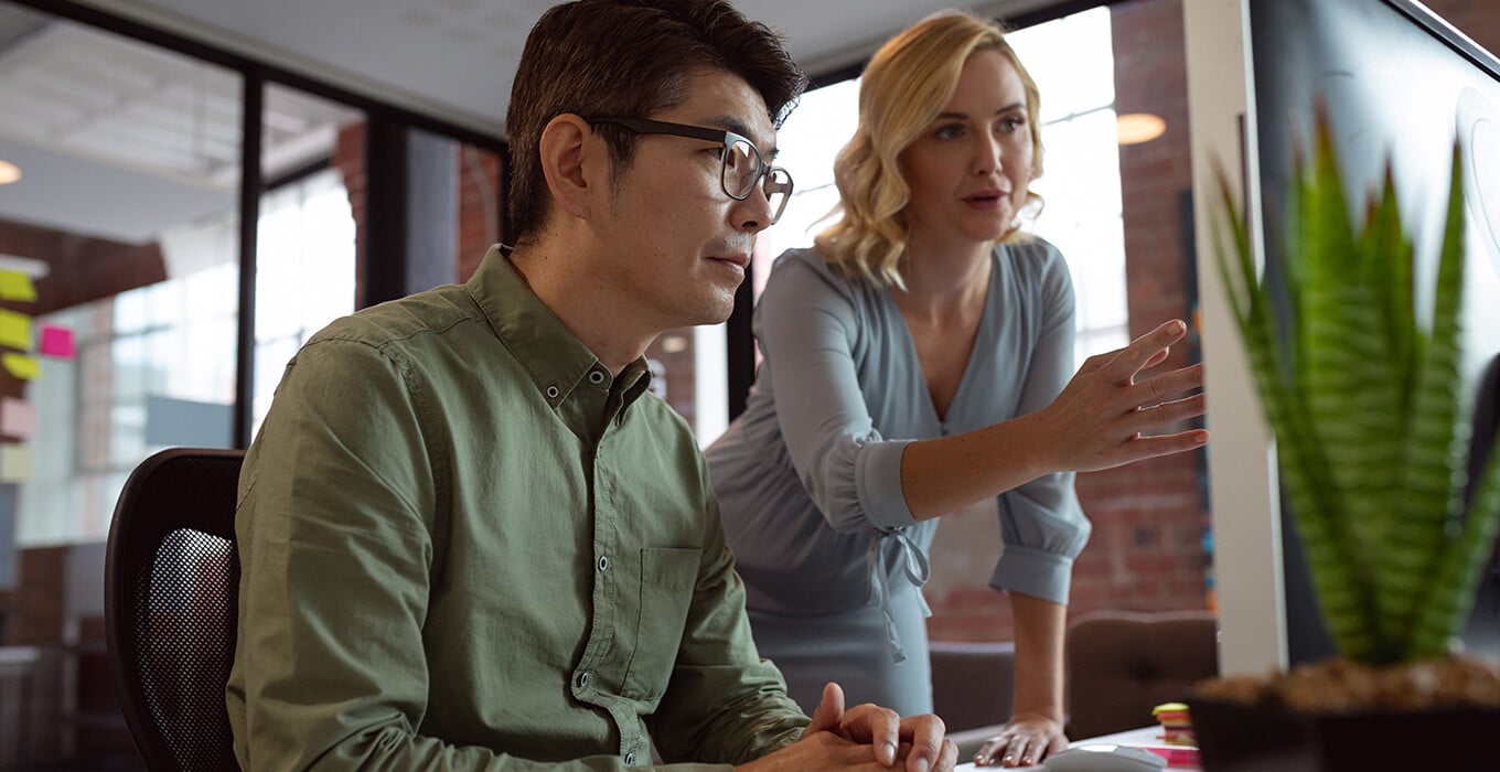 Two colleagues collaborating at a desk, with a woman pointing at a computer screen while a man in glasses closely observes